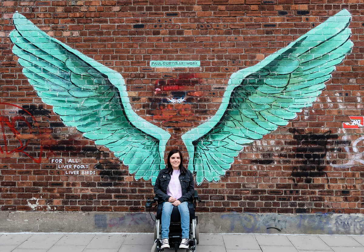 Emma sitting in her wheelchair against a brick wall. The wall has giant green angel wings spray painted on it by artist Paul Curtis.