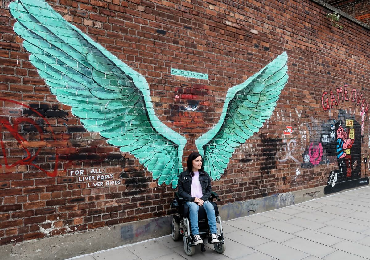 Emma sitting in her wheelchair against a brick wall. The wall has giant green angel wings spray painted on it by artist Paul Curtis. Emma has her wheelchair turned slightly away from the camera and she is looking off to the side. 