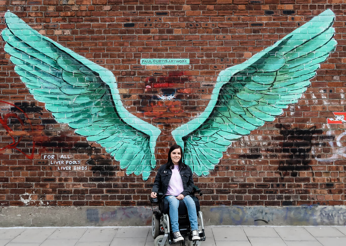 Emma sitting in her wheelchair against a brick wall. The wall has giant green angel wings spray painted on it by artist Paul Curtis.