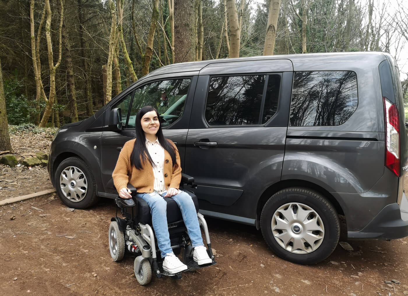 Emma, a power wheelchair user sitting in front of her wheelchair accessible vehicle. Emma is smiling and looking at the camera.