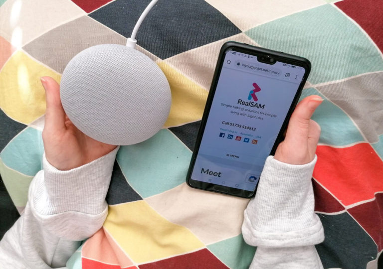 A view looking down at Emma's hands. One hand is holding the Google Nest Mini speaker and the other hand is holding her mobile and on the screen is the RealSAM website. The background is multicoloured.