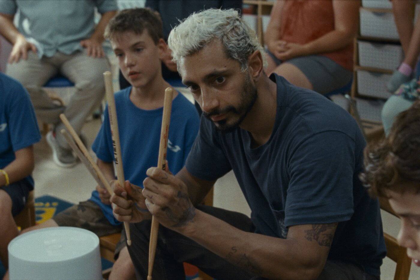 Riz Ahmed with bleached hair is sitting holding drum sticks. Next to him is young deaf students