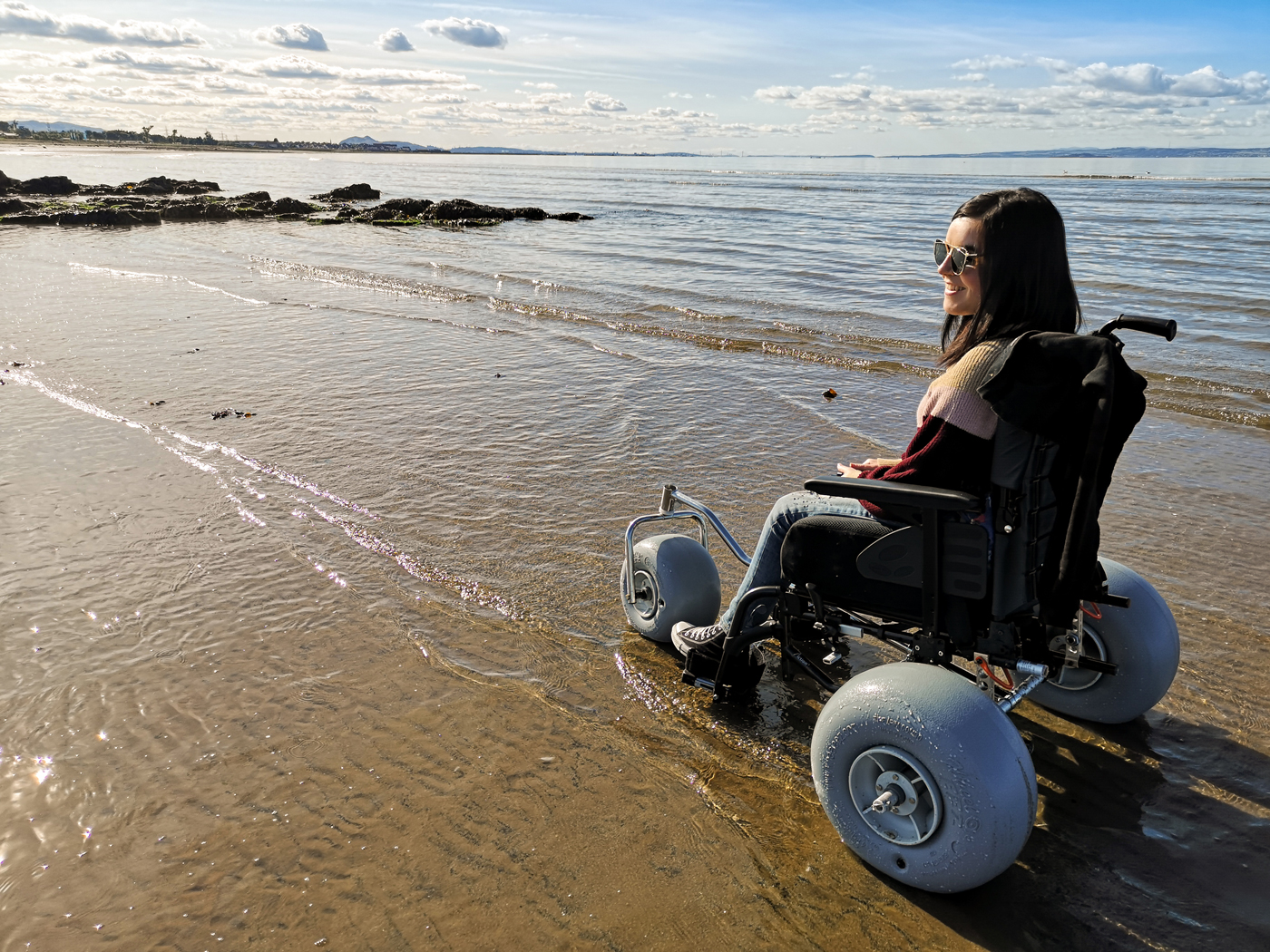 Emma sitting in a beach wheelchair on the beach at the edge of the water. Emma is looking out to the sea smiling.