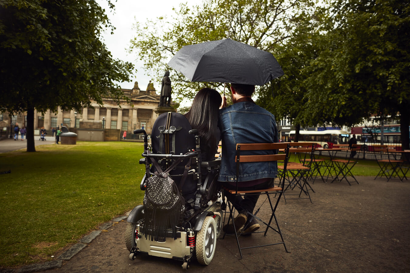 Emma and Allan sitting at a kiosk in Princes Garden, Edinburgh. They are sitting at a table with their backs to the camera. Allan is holding an umbrella over them.