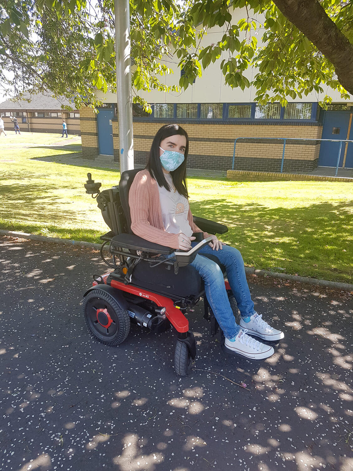 Emma, a white woman sitting in her powered wheelchair outside next to a grassy area and tree. Emma is wearing a peach cardigan, skinny jeans and white converse shoes. She has a face mask covering her nose and mouth.