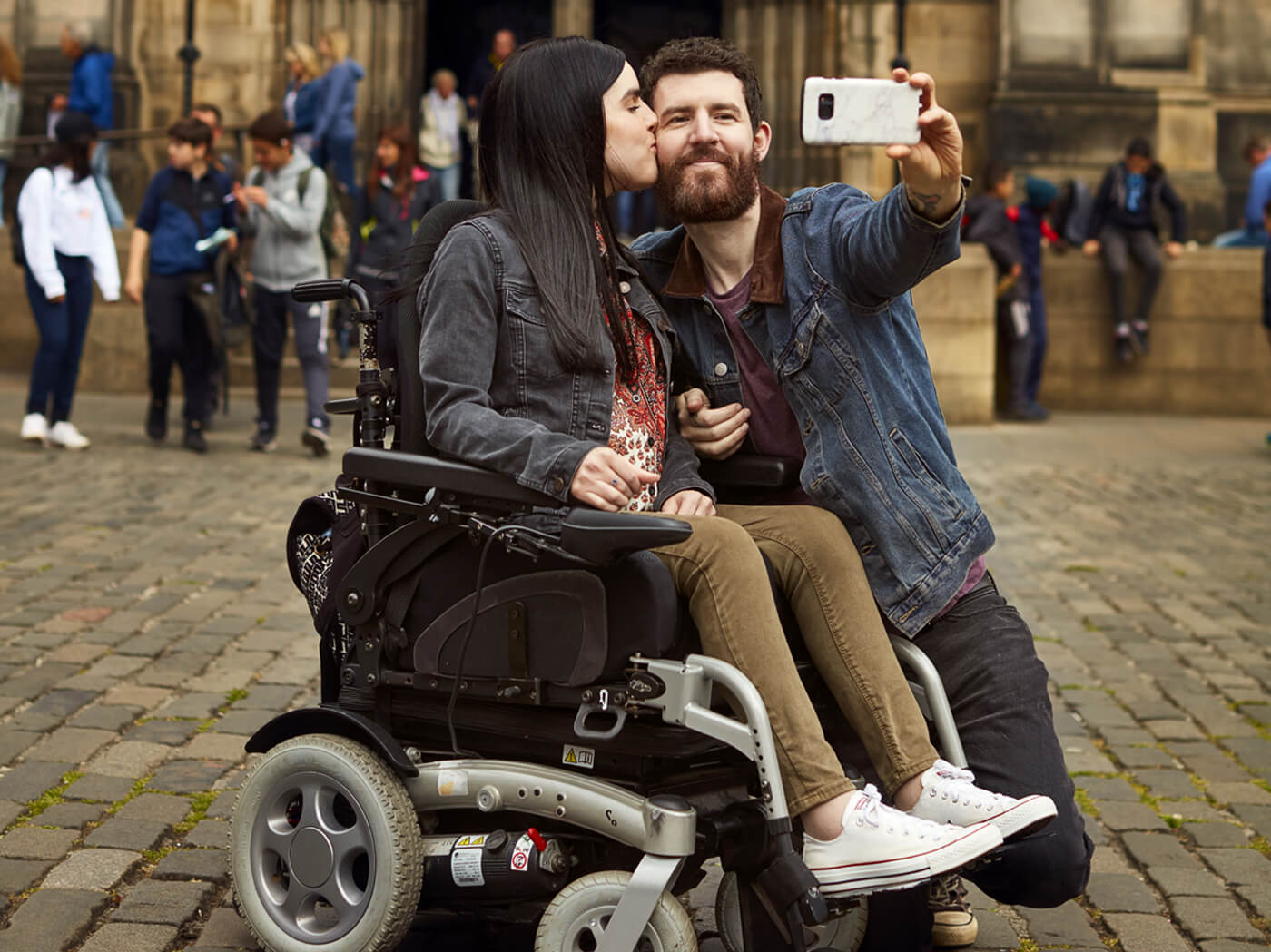 Emma in her powered wheelchair with her boyfriend kneeling beside her holding a phone up while taking a selfie of them together. Emma is giving him a kiss on the cheek. They are outside a Cathedral in Edinburgh.