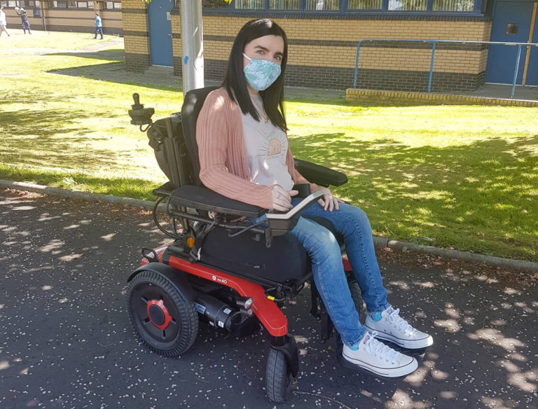 Emma, a white woman sitting in her powered wheelchair outside next to a grassy area and tree. Emma is wearing a peach cardigan, skinny jeans and white converse shoes. She has a face mask covering her nose and mouth.