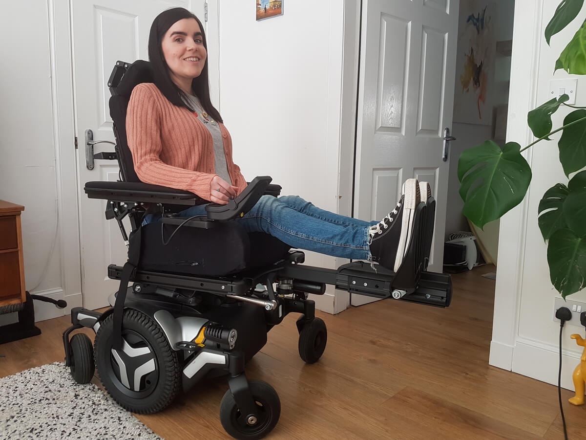 Emma, a wheelchair user is sitting in a black and silver Permobil wheelchair. She has her legs raised on the footplates. She is wearing a peach cardigan, grey t-shirt, blue skinny jeans and black converse shoes.
