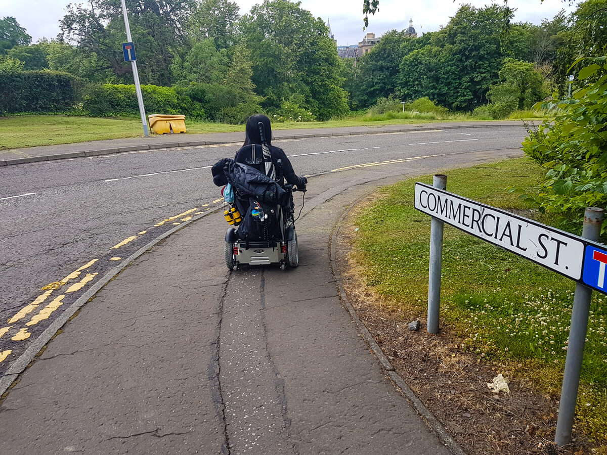 Emma, driving her wheelchair on a path down towards a park. A street sign called 'Commerical St.' is on the right of the photo.
