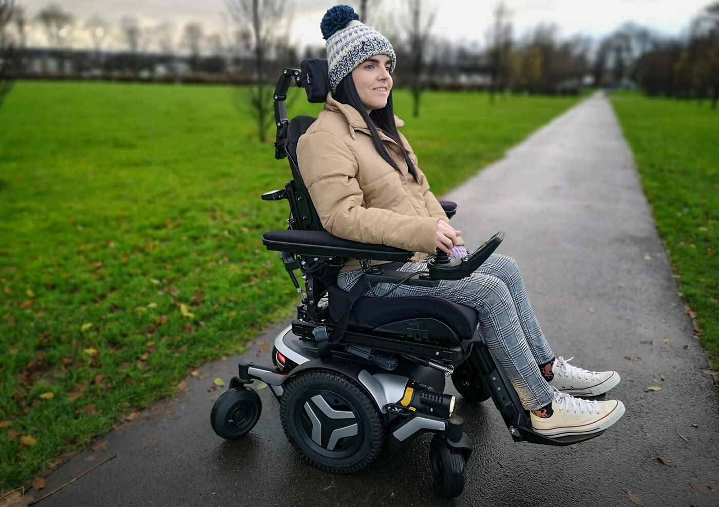 Emma, a wheelchair user sitting in a black and silver Permobil M3 wheelchair in a park. She is wearing a woolly hat, puffy jacket, black and white check trousers and white converse shoes. Emma is smiling.