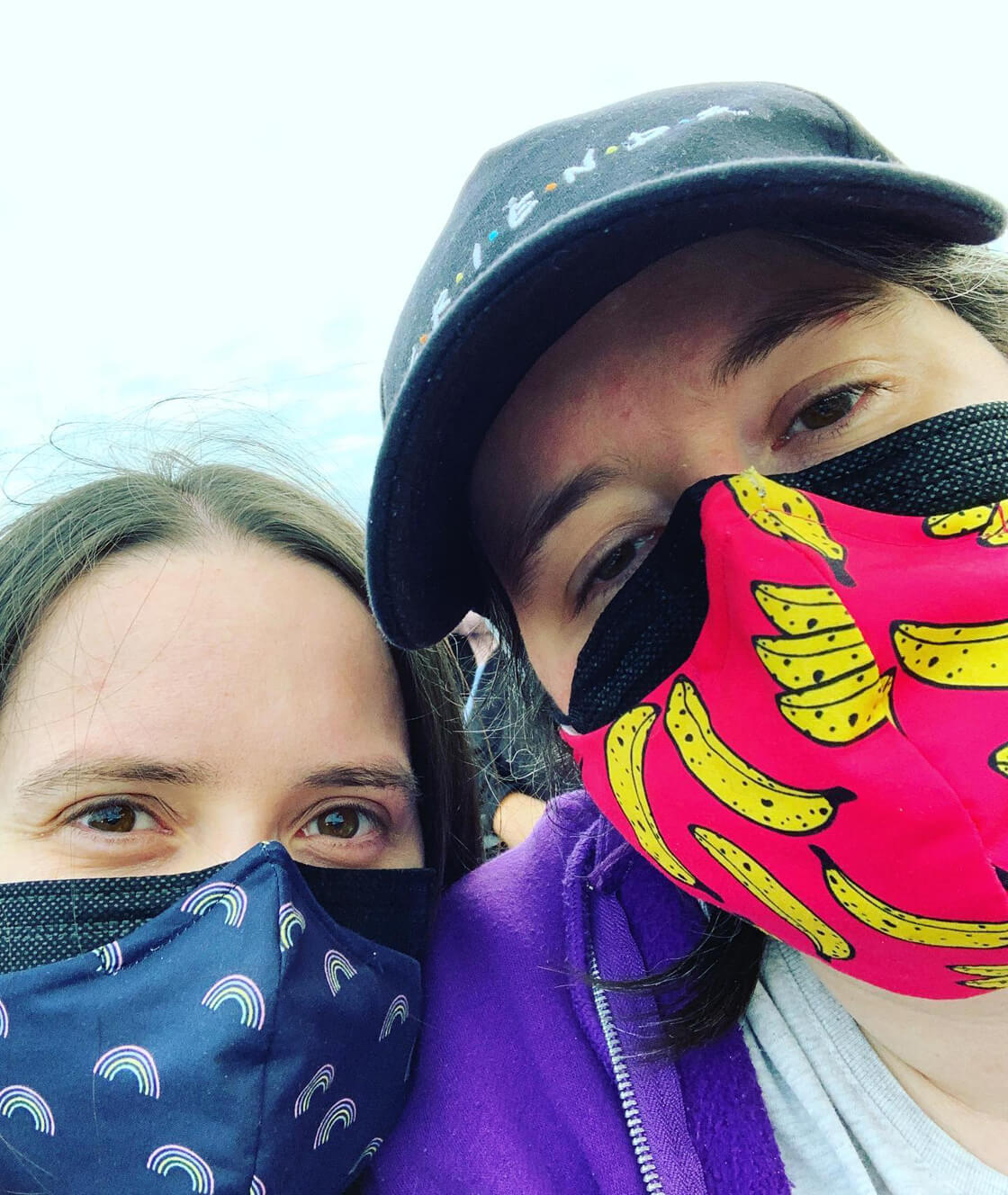 A selfie from the shoulders up of Sarah and karine. They are both wearing face coverings. Karine is wearing a black Friends hat.