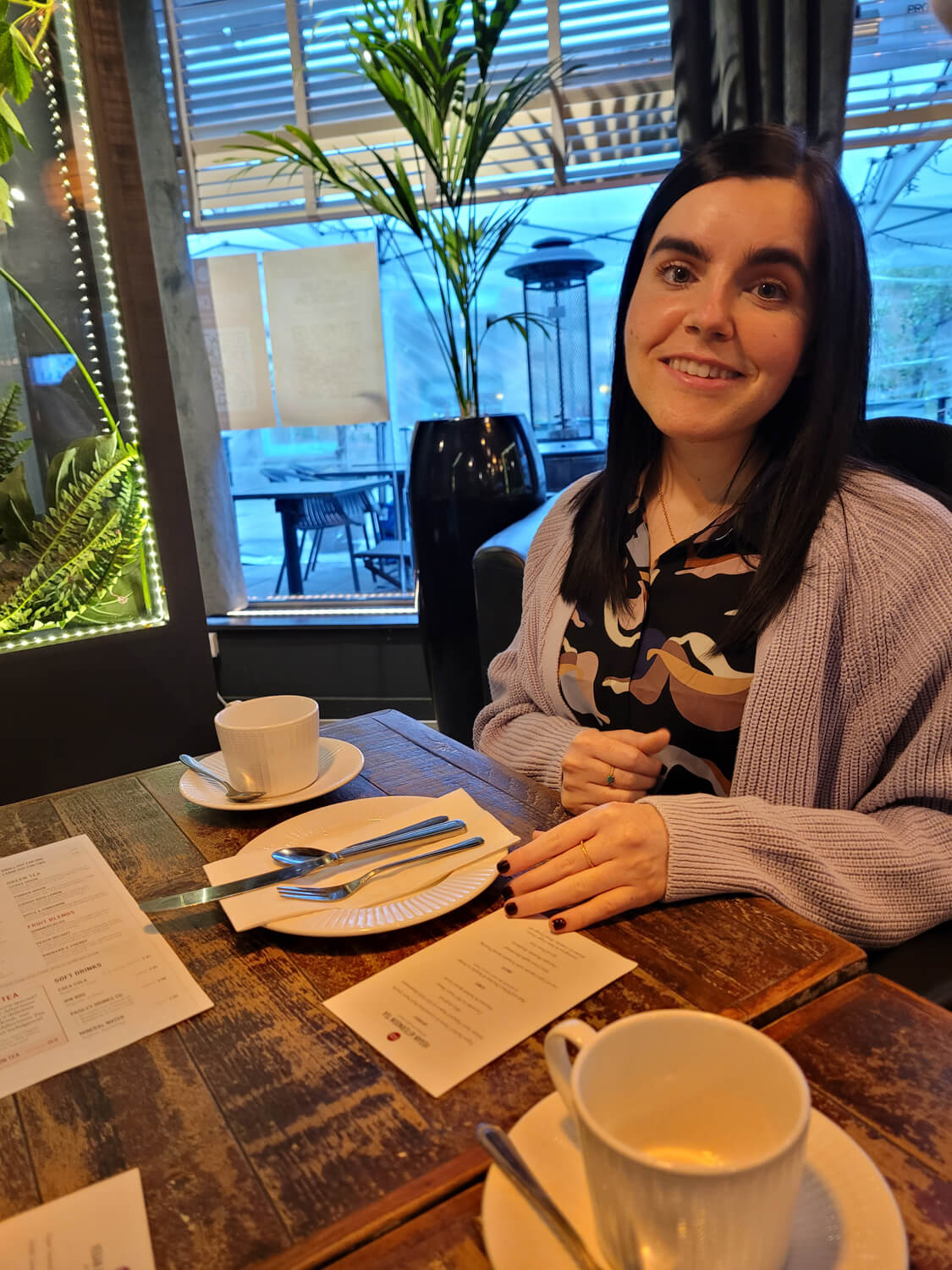 Emma sitting at the table in the restaurant. She is wearing a purple cardigan with a black blouse with different patterns and shapes.