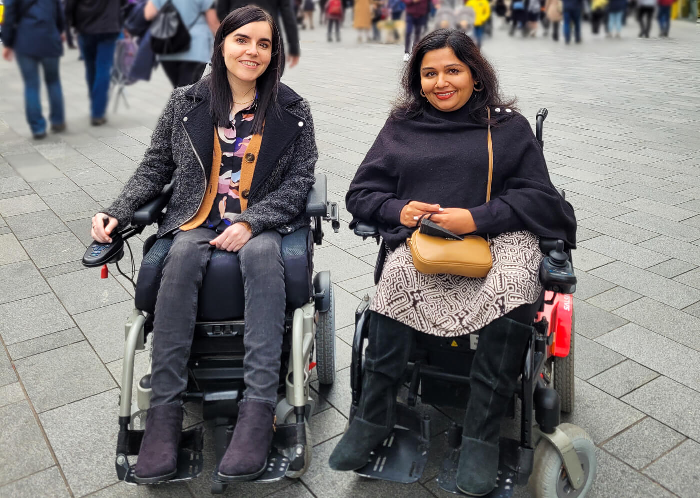 Emma and Tanvi are female power wheelchair users. They are sitting beside each other in a London city centre street. They both have dark hair and smiling at the camera.