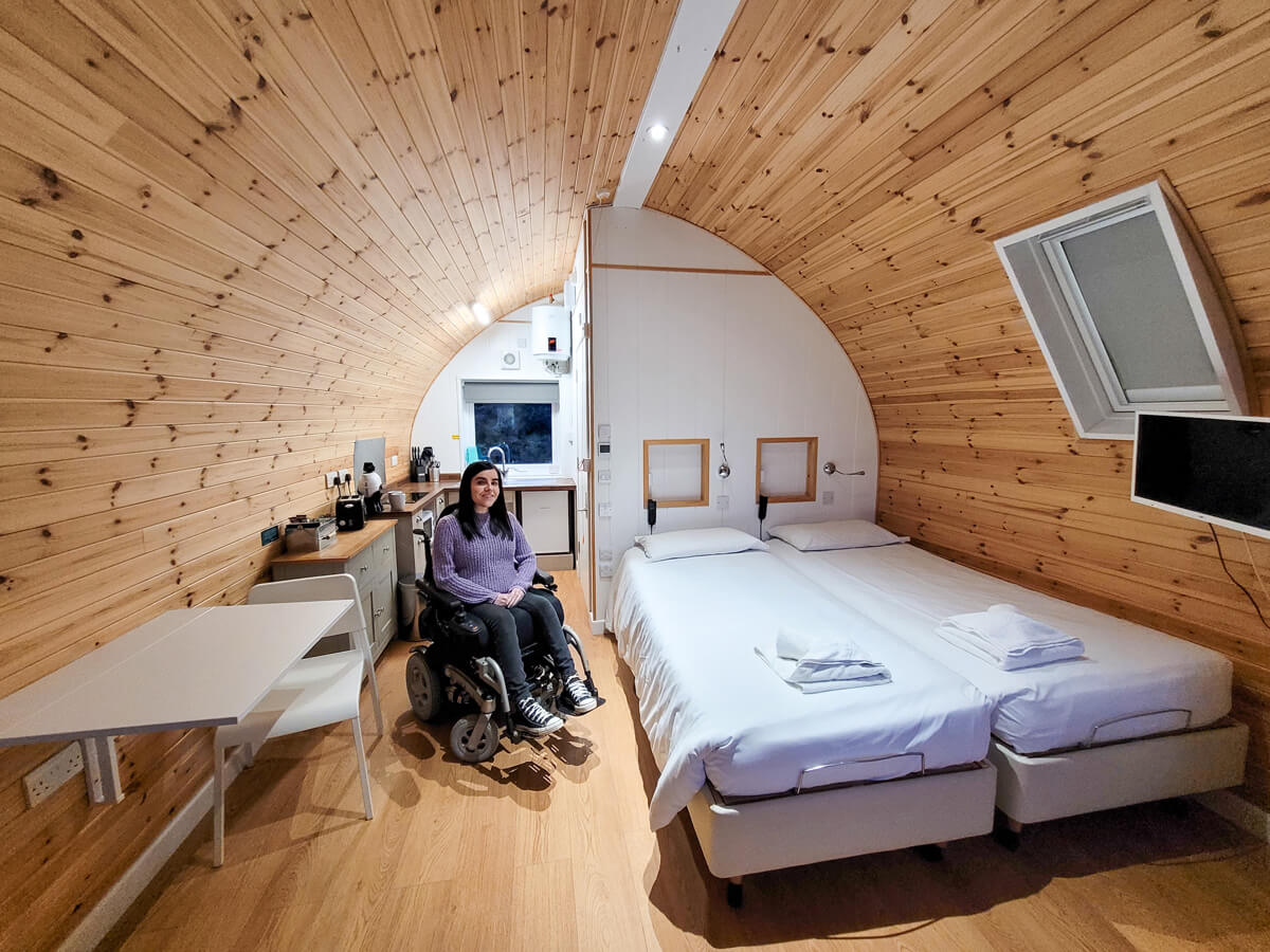 Emma, a female power wheelchair user is sitting in the accessible glamping pod next to the profiling bed. She is smiling at the camera. The photo shows the entire pod including bed, kitchen and dining area.