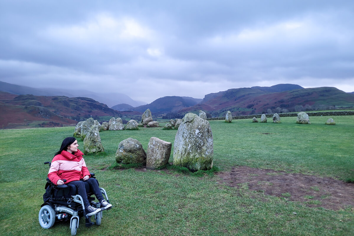 Emma is sitting her wheelchair while looking away to the left. She is at Castlerigg Stone Circle. The clouds are grey and look very dramatic. Emma is surrounded by mountains.