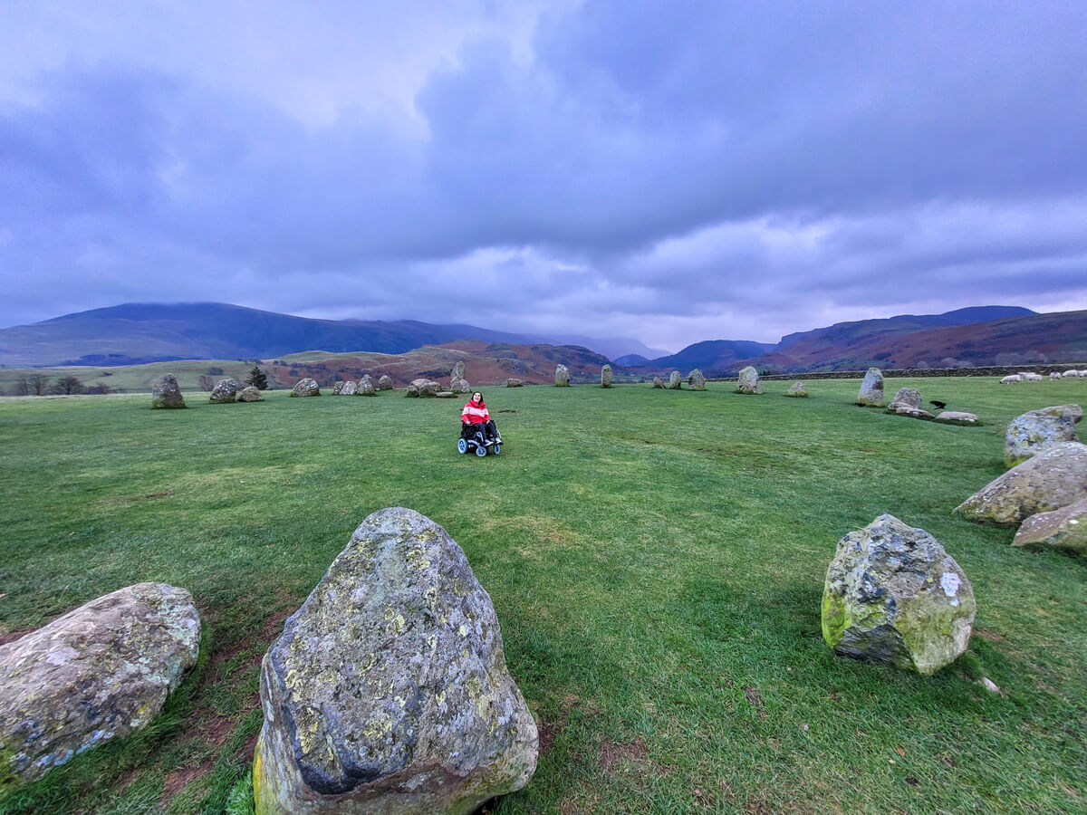 Emma is sitting her wheelchair in the middle of Castlerigg Stone Circle. The clouds are grey and look very dramatic. Emma is surrounded by mountains.