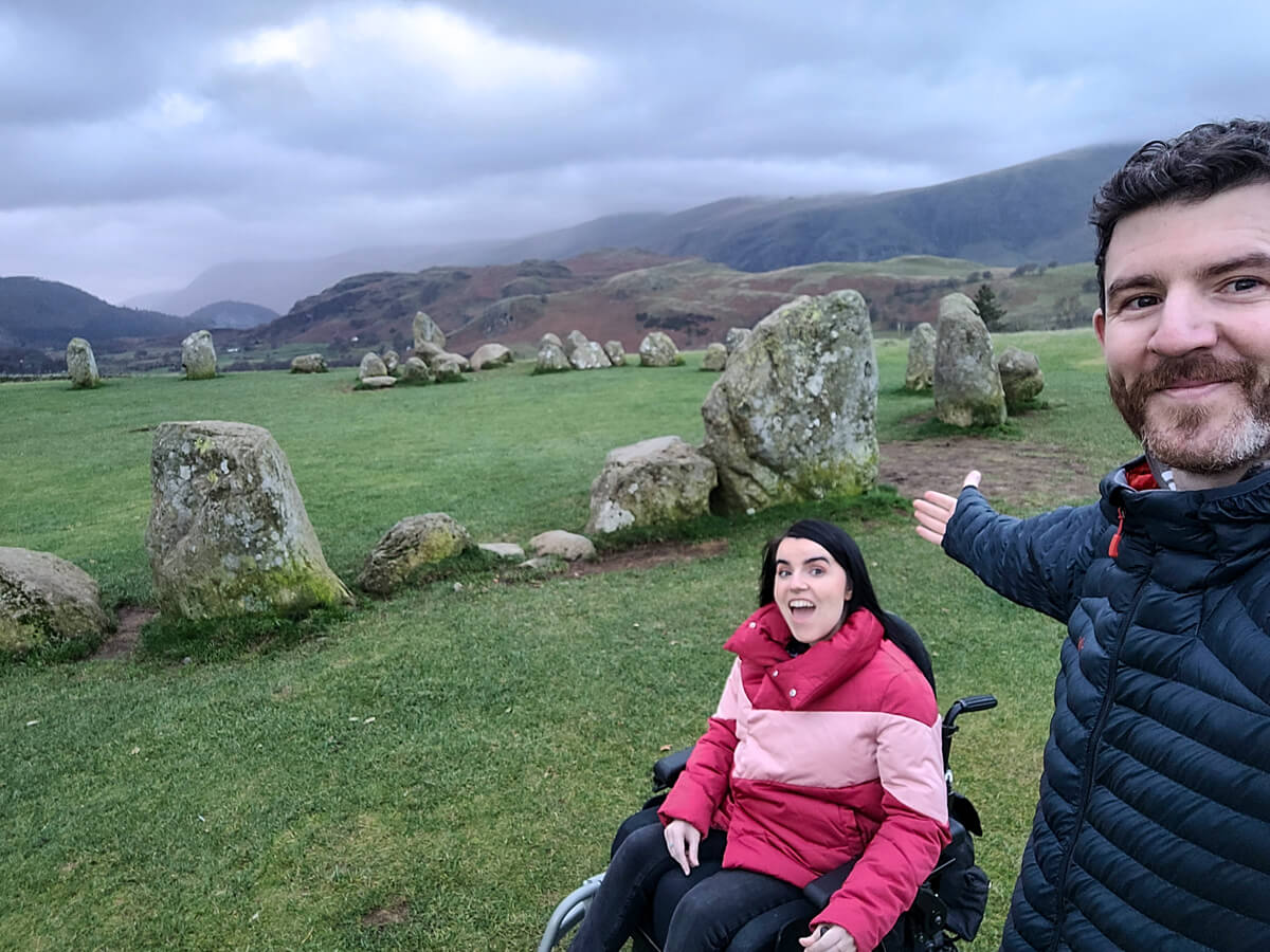 Emma and Allan on a hill at the top of Castlerigg Stone Circle. Emma is smiling and Allan has his right arm up while also smiling. The clouds are grey and look very dramatic. They are surrounded by mountains.