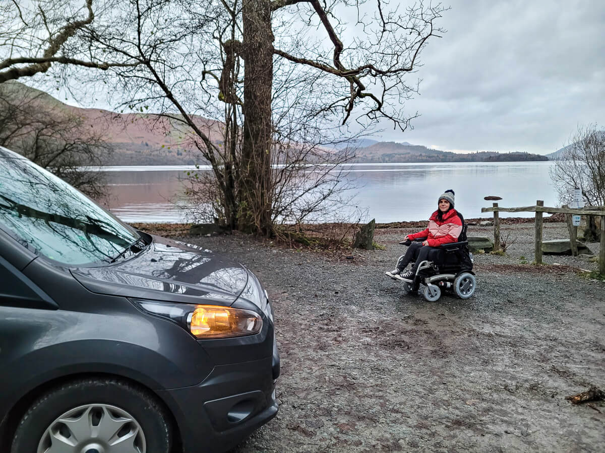 Emma is sitting in her powered wheelchair wearing a woolly hat and red puffy jacket and black jeans. Derwentwater is behind her as a beautiful backdrop.