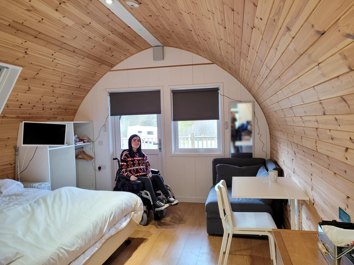 Emma wearing a Christmas jumper and smiling at the camera. She is sitting next to the door inside the glamping pod. The end of the bed is in view as well as the dining table, TV and sofa bed.