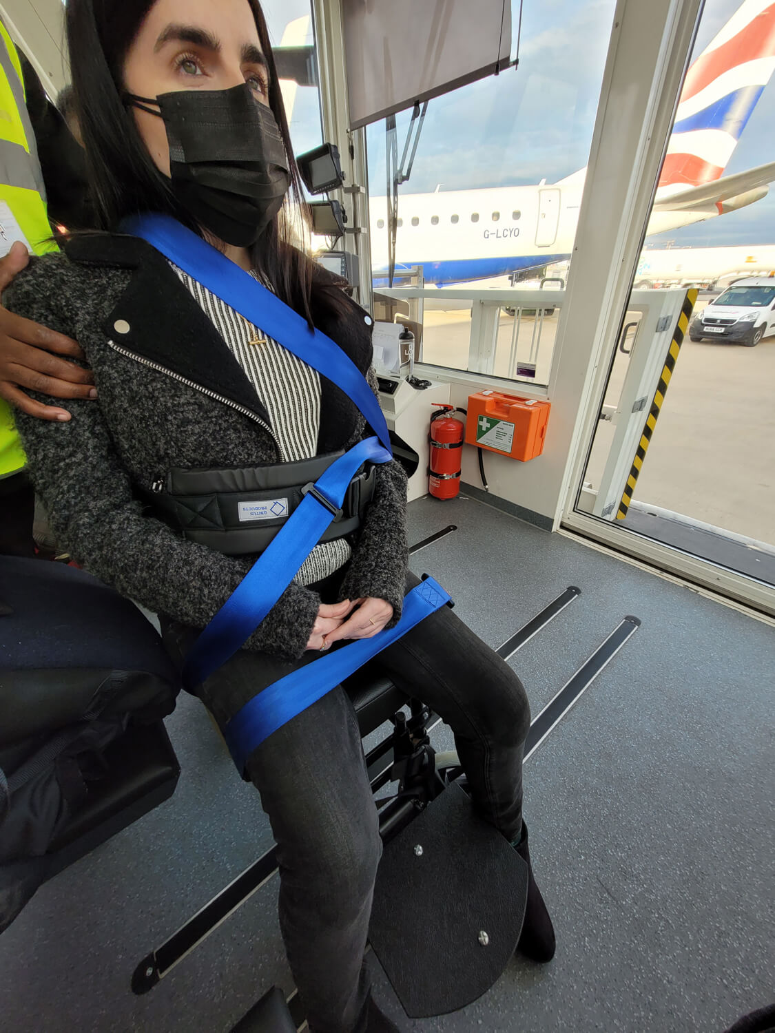 Emma strapped into an airport aisle chair inside the ambulift. The British Airways flag on the plane tail can be seen through the ambulift window. Emma is looking solemly straight ahead. Her feet are dangling at the side of the footplate.