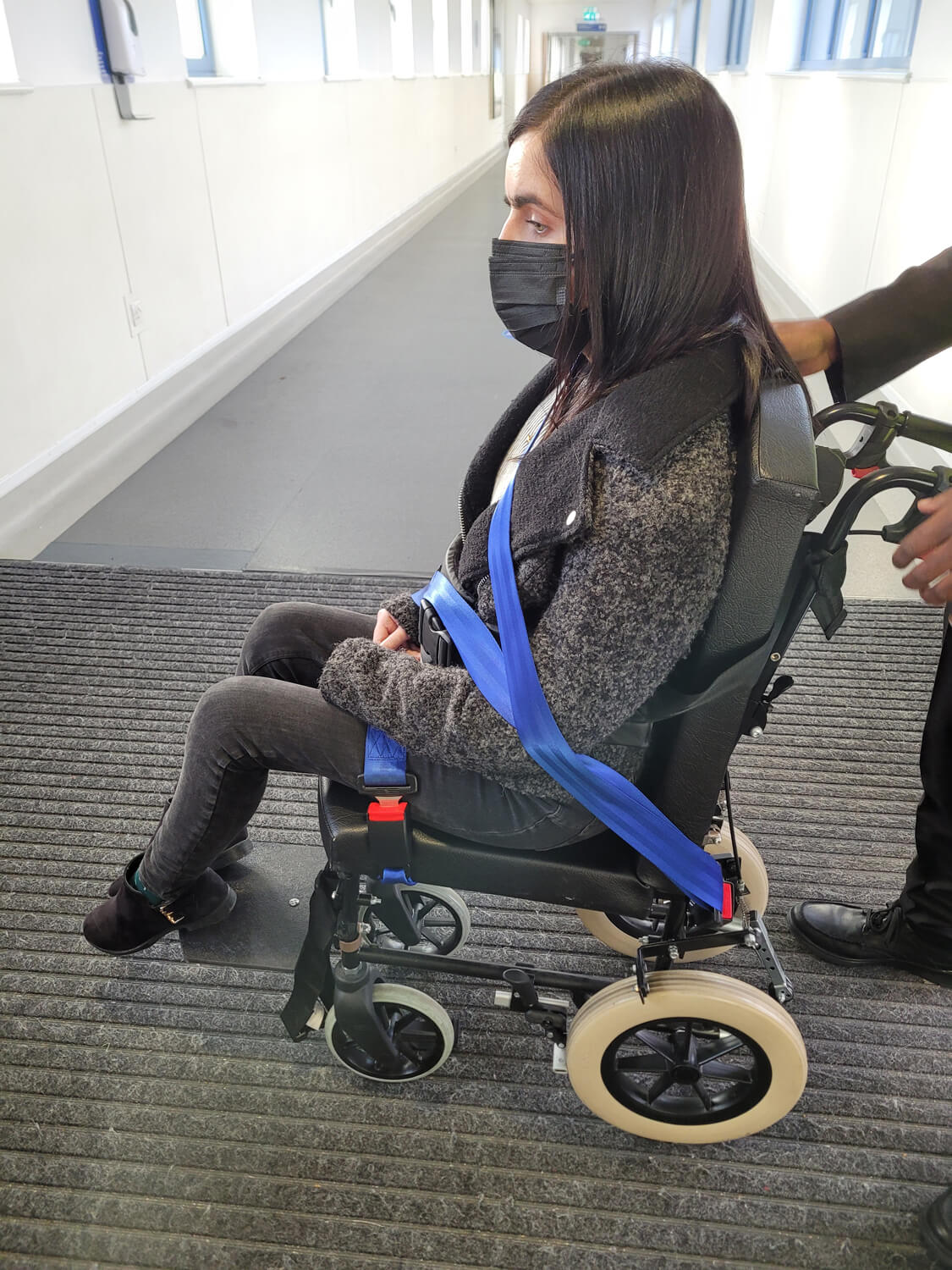 A side view of Emma strapped into an aisle chair at London City Airport. The heel of Emma's boots are on the footplate while her feet are pointing directly down to the ground. She is wearing a black face mask, black jacket, black jeans and black boots. The special assistance agent has his hand on her shoulder.