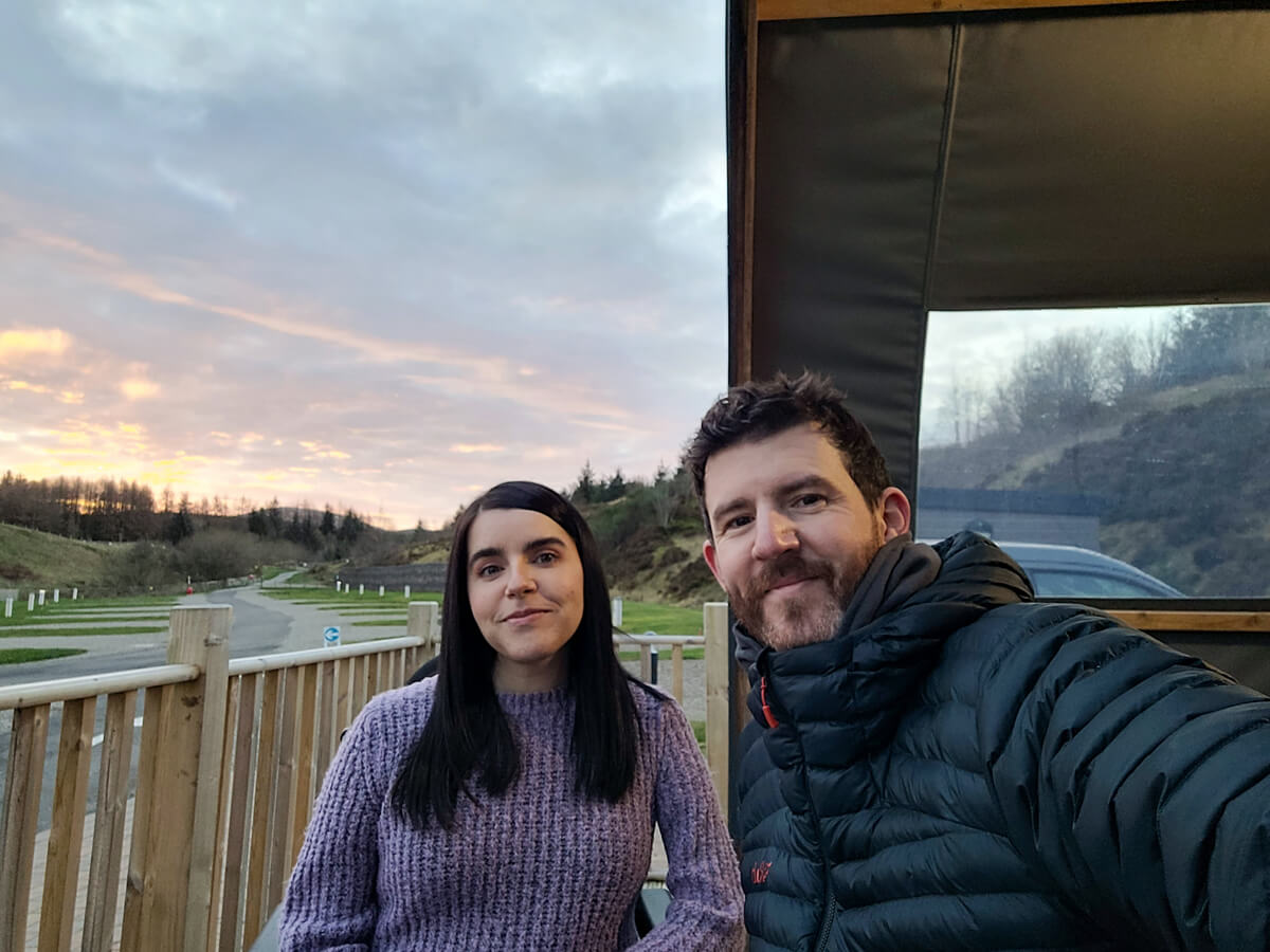 Allan and Emma from the waist up. They are on the deck area of the glamping pod at Troutbeck Head. The sun is setting behind them.
