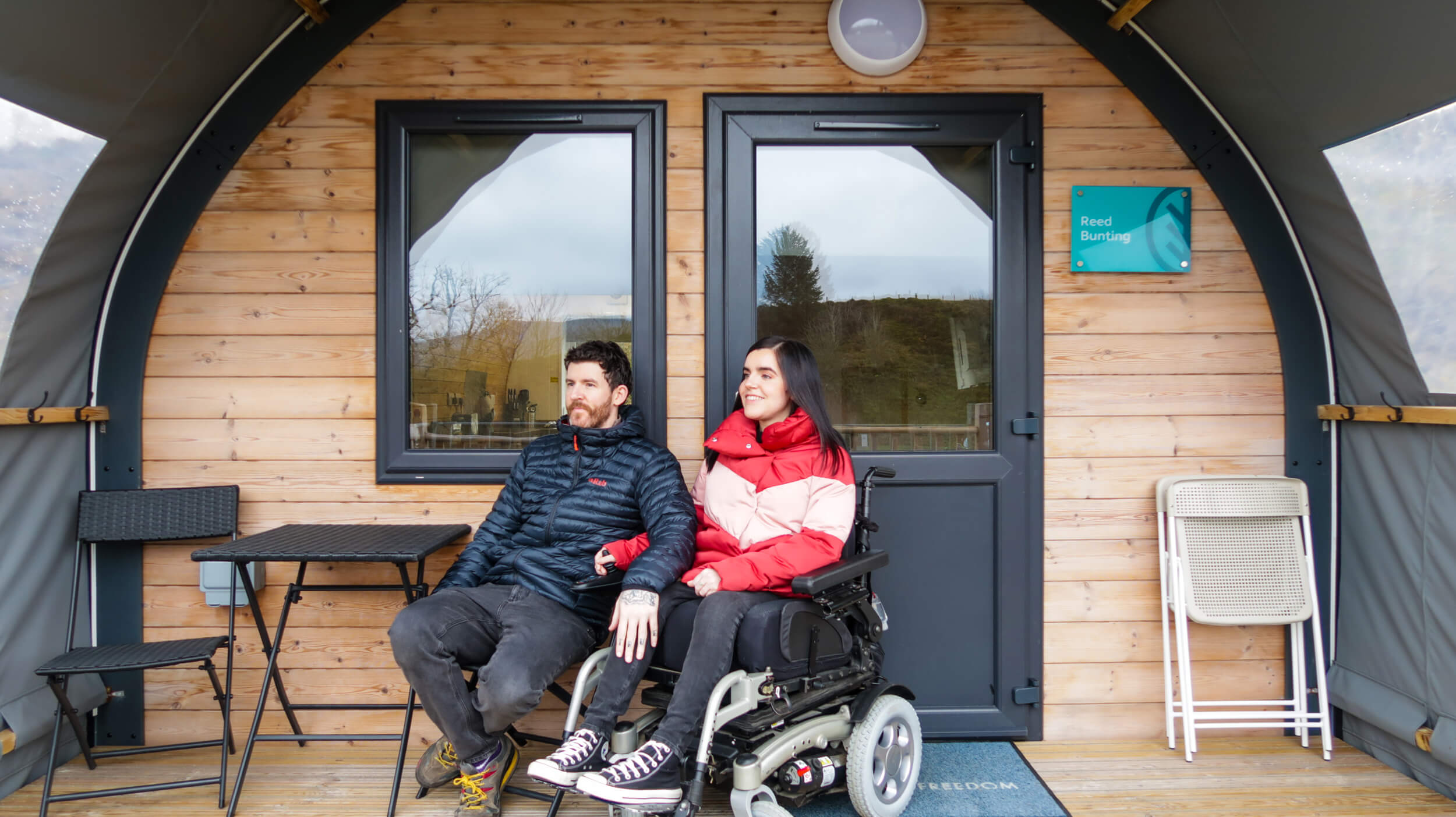 Emma and Allan sitting on the wooden deck under a sheltered canopy roof. The door to the accessible glamping pod is directly behind them. Emma is wearing a red puffy jacket, black jeans and black converse. Allan is wearing a dark blue puffy jacket, black jeans and walking shoes. They are both smiling.