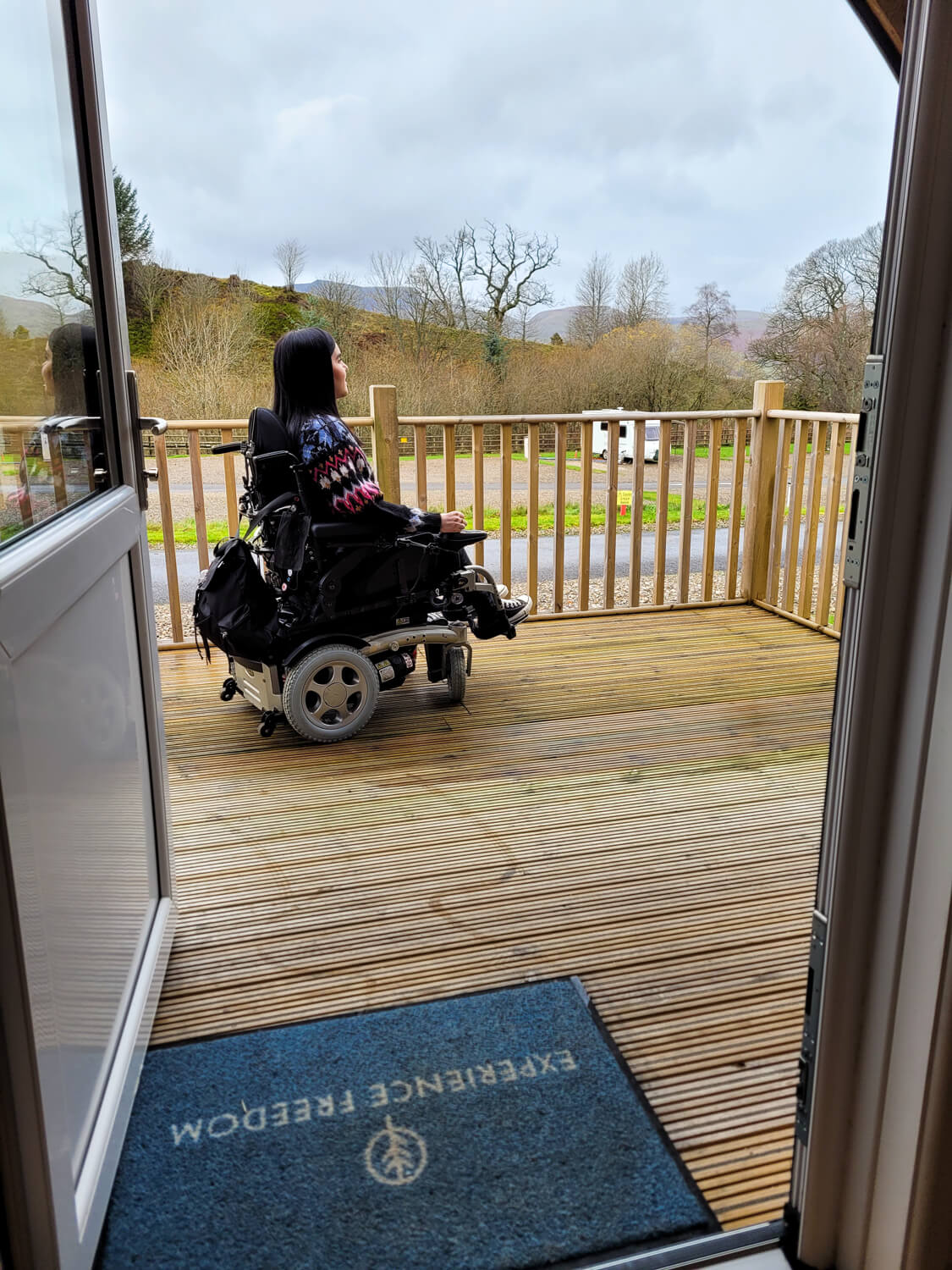 A view from inside the omnipod, looking out to Emma sitting on the decking in her power wheelchair. Emma is looking out at the view of the site with trees and hill. The door mat is in view at the bottom of the photo, it has the Experience Freedom logo.