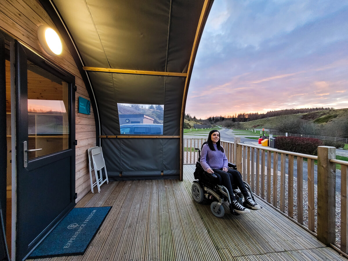 A side view of Emma sitting on the wooden deck under a sheltered canopy roof outside the accessible glamping pod at Troutbeck Head. She is wearing a purple jumper, black jeans and black converse. Emma is smiling. A purple, pink and orange sunset is behind Emma.