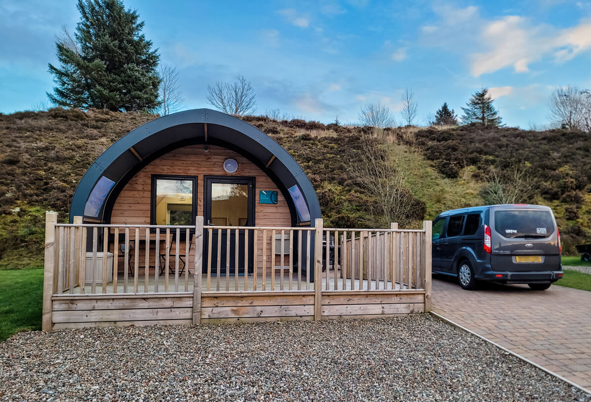Wheelchair accessible glamping pod at Troutbeck Head. A car is parked on the bricked driveway next to the pod. Behind the pod is shrubs and trees. The sky is blue with white wispy clouds.