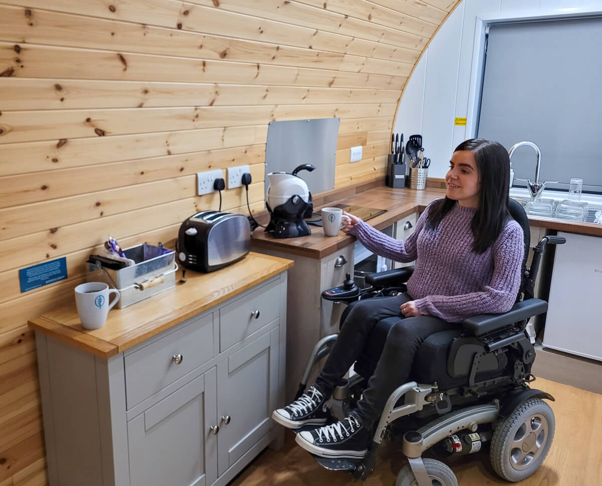 Emma a power wheelchair user is in the kitchen of the wheelchair accessible glamping pod at Troutbeck Head. She is wearing a purple knit jumper, black jeans and black concerse. She has dark long hair and is smiling. Her right hand is holding a mug which is on the worktop.