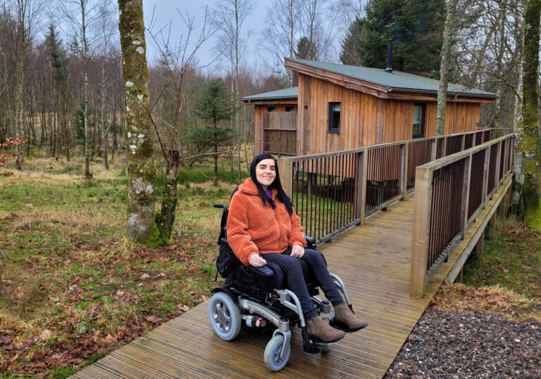 Emma is sitting in her power wheelchair on a wooden ramp leading to her accessible treehouse. Emma is wearing an orange fluffy jacket, black jeans and khaki boots. Her dark hair is long and styled down. She is smiling at the camera. The beautiful wooden treehouse is behind her and is surrounded by trees.