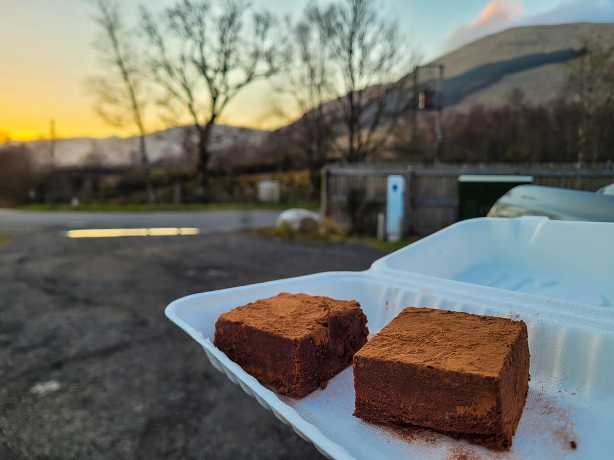 Two vegan chocolate brownies in a takeaway box with mountains as a view in the background.