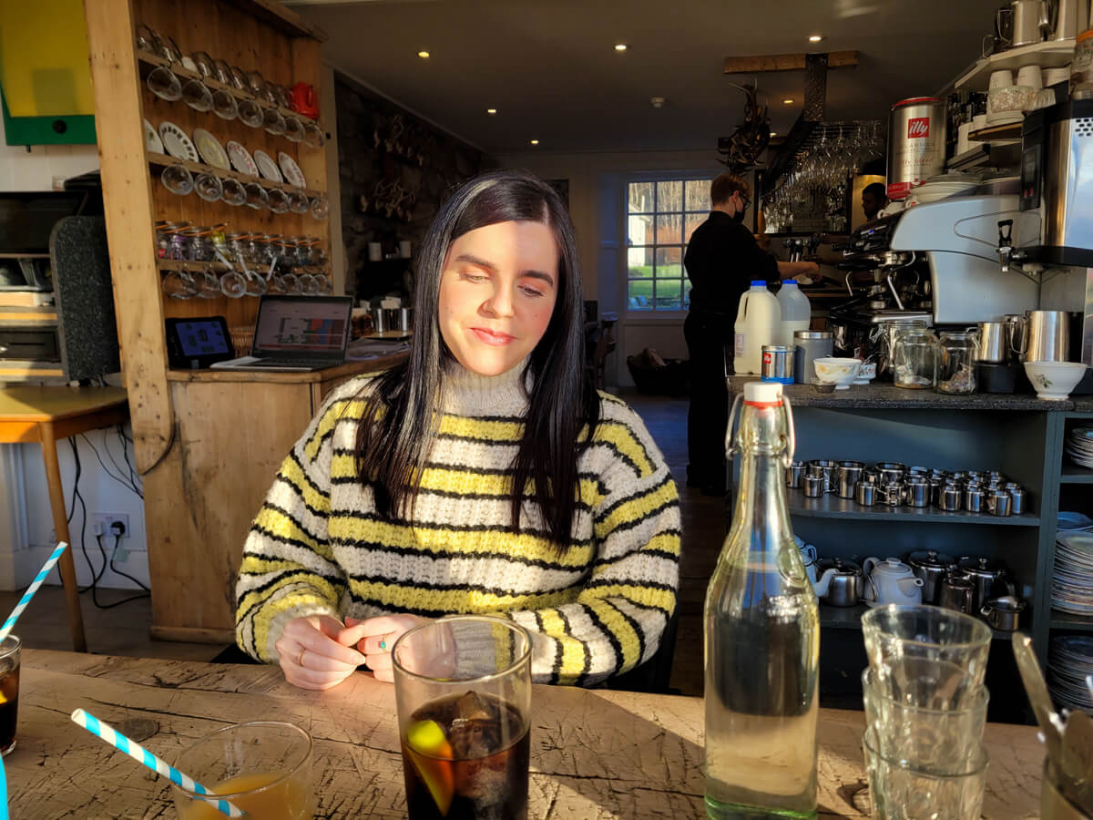 Emma sat a table in Mhor 84 restaurant. She is smiling and looking down at the table. Sun is shining on her face.