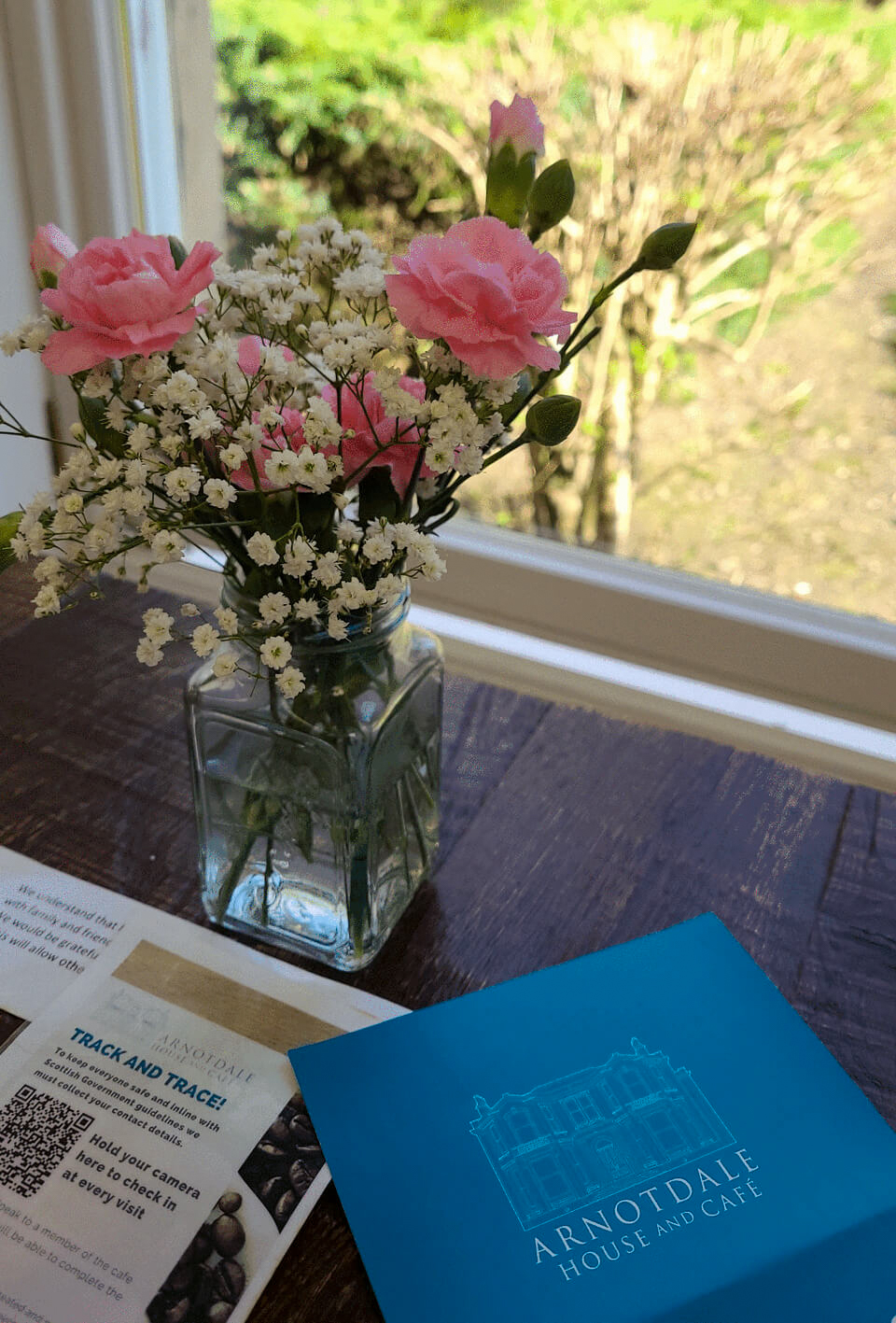 A glass vase with pink and white flowers sat on a table next to a window with greenery outside. On the table is a blue menu for Arnotdale House and Cafe.