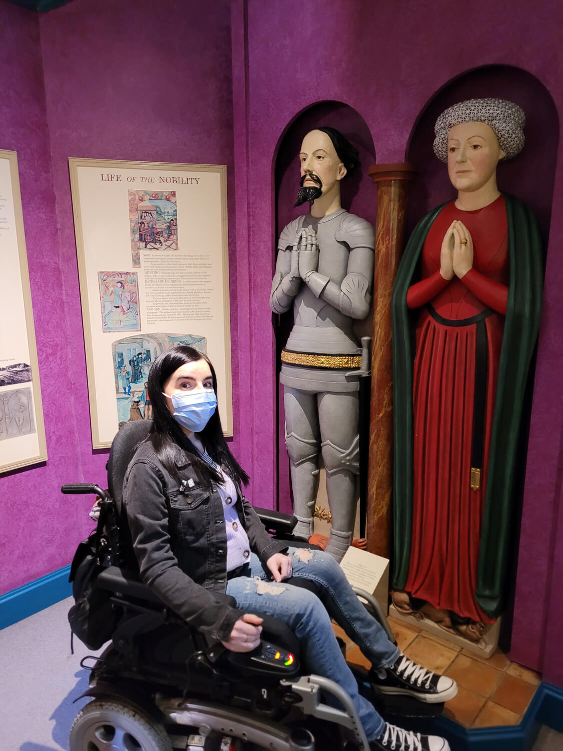 Emma sat in her wheelchair wearing a face mask. She is next to a display of medieval people.