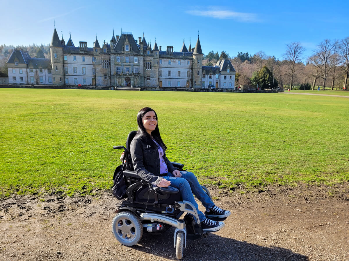 Emma sat in her wheelchair with the stunning Callendar House behind her. Emma is smiling at the camera and the sun is shining.