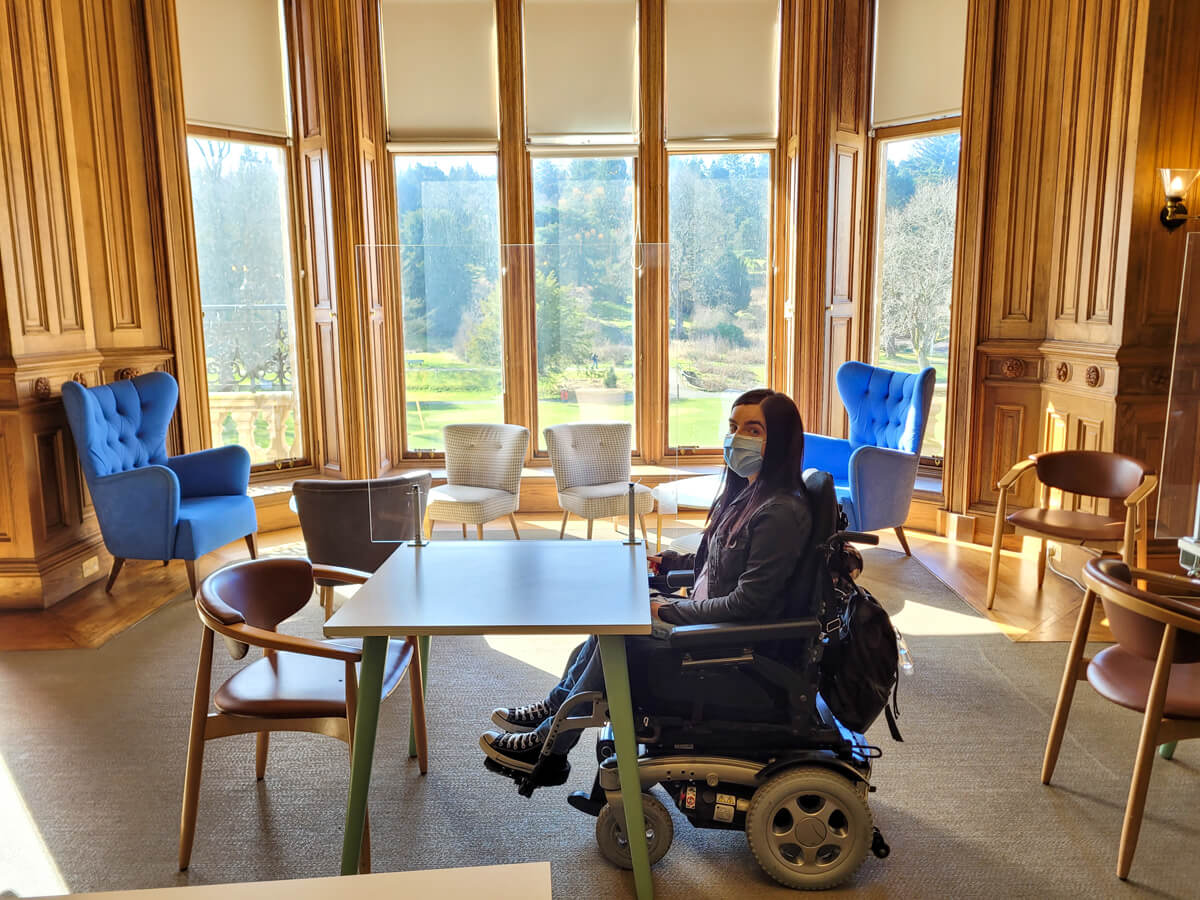 Emma sat in her wheelchair at a table in the Callender House tearoom. The large ceiling to floor windows with a view out to the lush green park is behind Emma.