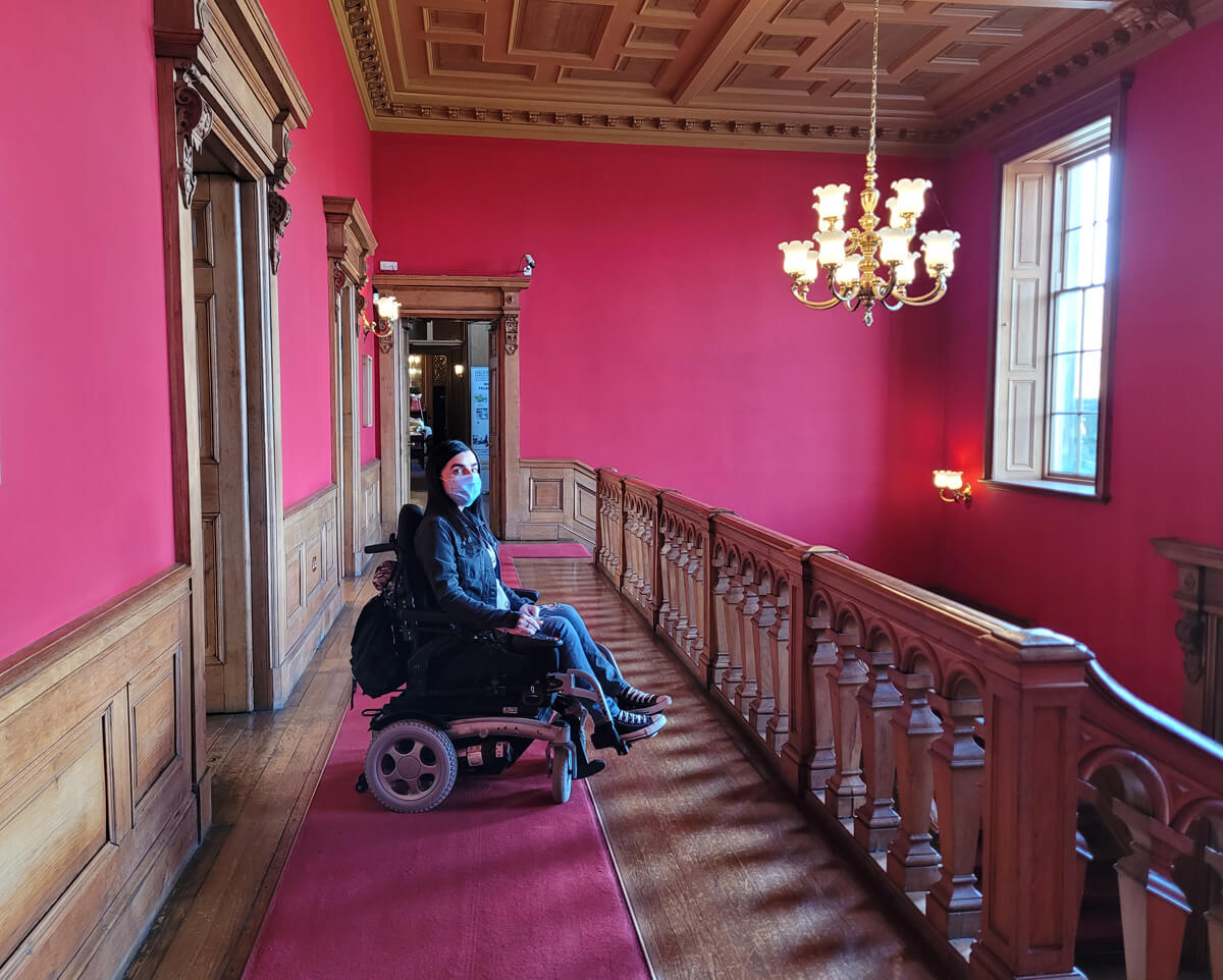 Emma sat in her wheelchair inside Callendar House. She is in the grand hallway with red walls, a gold chandelier and dark wood.