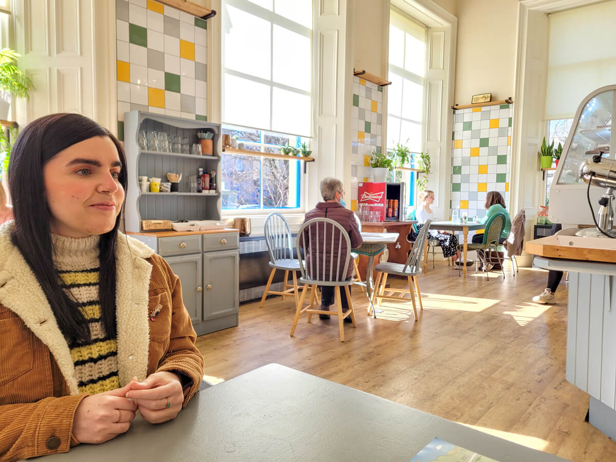 Emma is sitting a table in Smith Cafe. She is looking off to the side. The cafe is bright with white, yellow and green tiles. Plants are displayed on the window ledge. Her dark hair is long and she is wearing a stripy jumper and mustard cord jacket.
