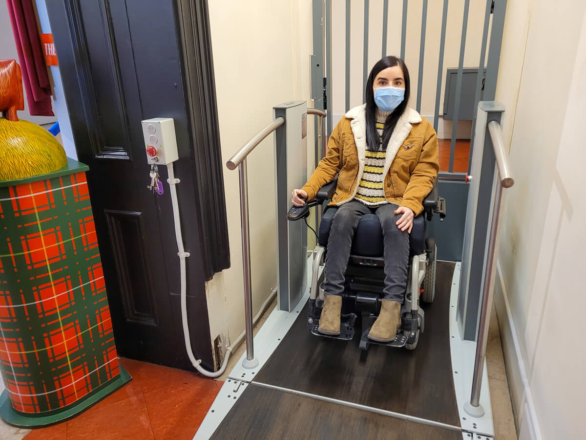 Emma sat in her power wheelchair on a wheelchair lift in the Smith Art Gallery and Museum. She is wearing a face mask, mustard cord jacket, black skinny jeans and khaki green boots.