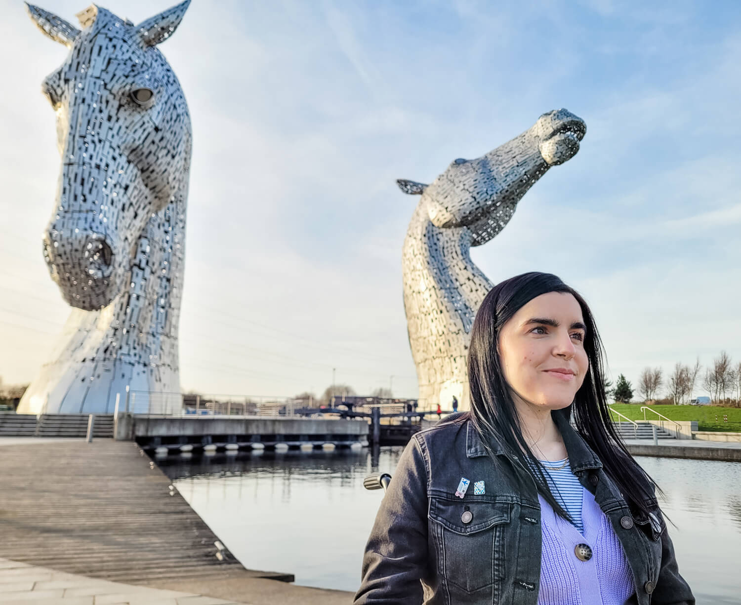 A shot of Emma from the waist up with the giant Kelpies behind her. Emma is smiling and looking off to the side of the camera. It is a sunny day and the Kelpies are shining.