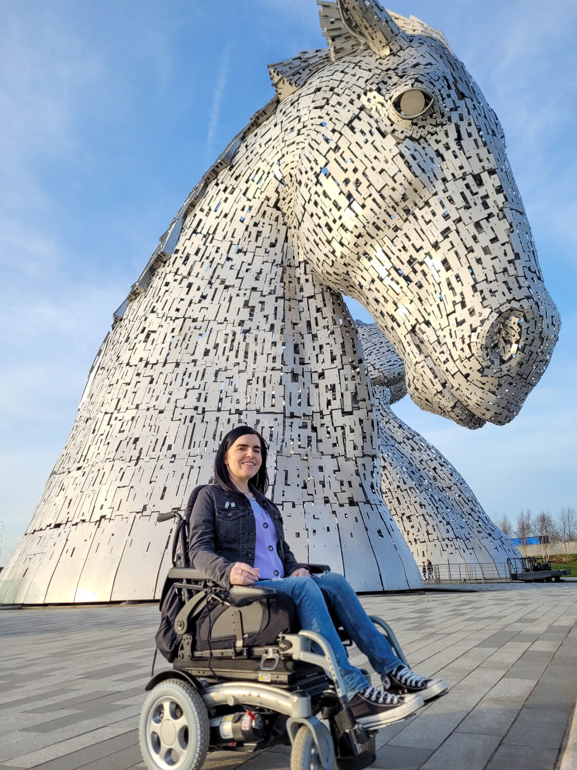 A full shot of Emma sat in her power wheelchair in front of the Kelpies in Falkirk. Emma is smiling at the camera.