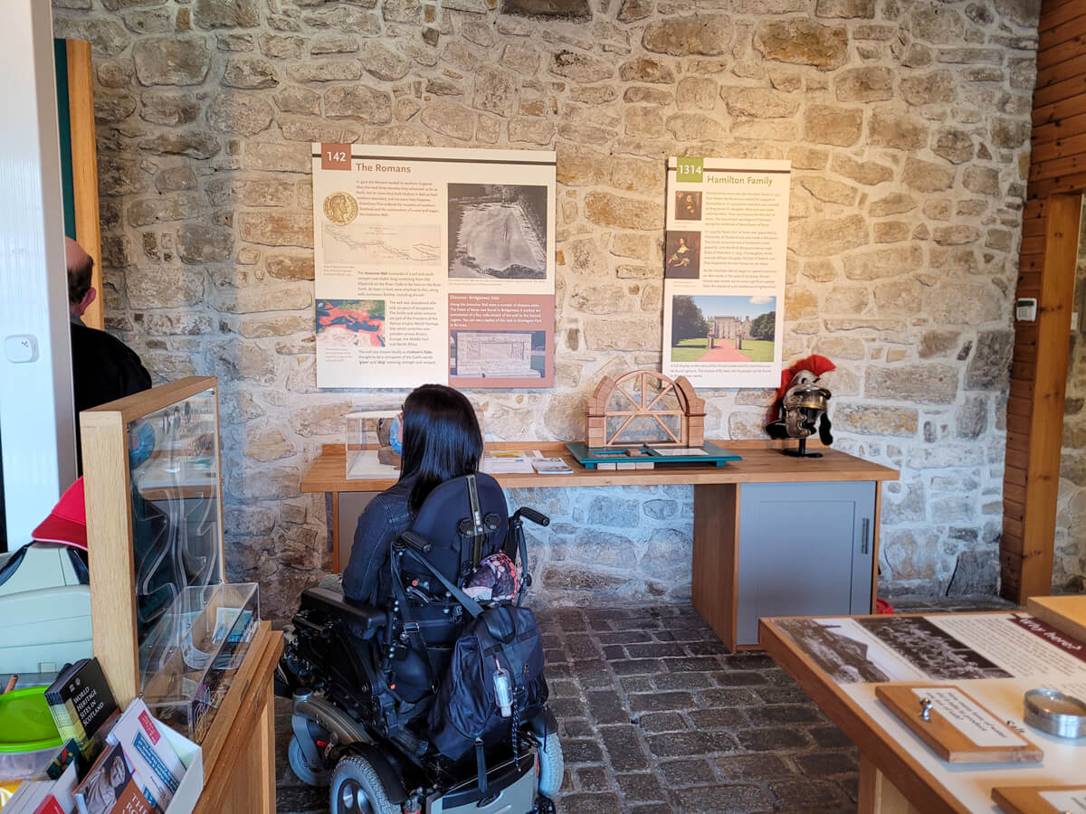 Emma sat in her wheelchair inside Kinneil Museum. She is looking at the information displays hanging on the wall.