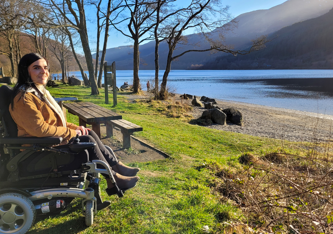 Emma sat in her power wheelchair on the grass next to loch Lubnaig. Emma is smiling at the camera.