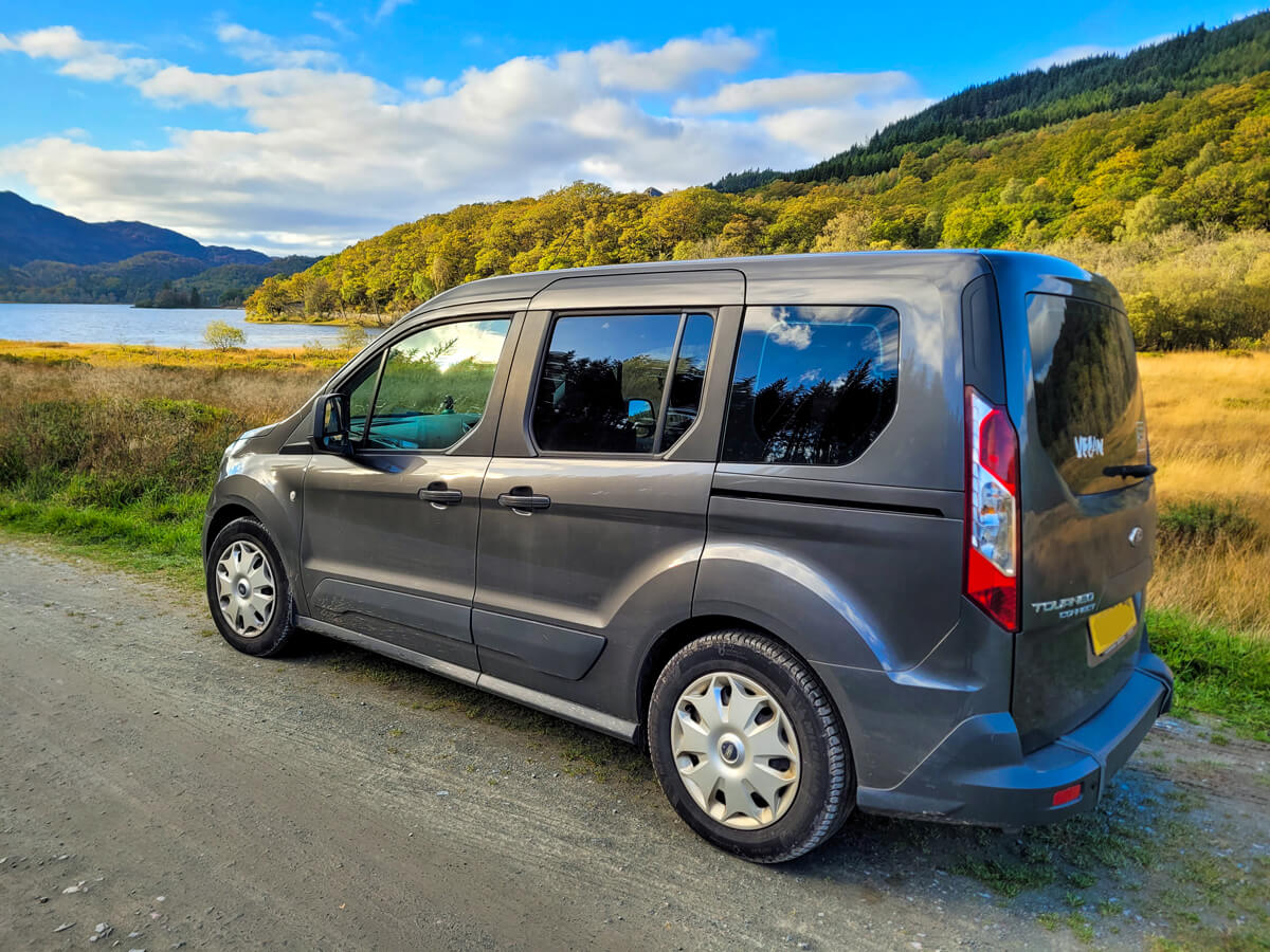 Ford Tourneo WAV parked up on the side of the road. There is a loch and mountains behind the car.
