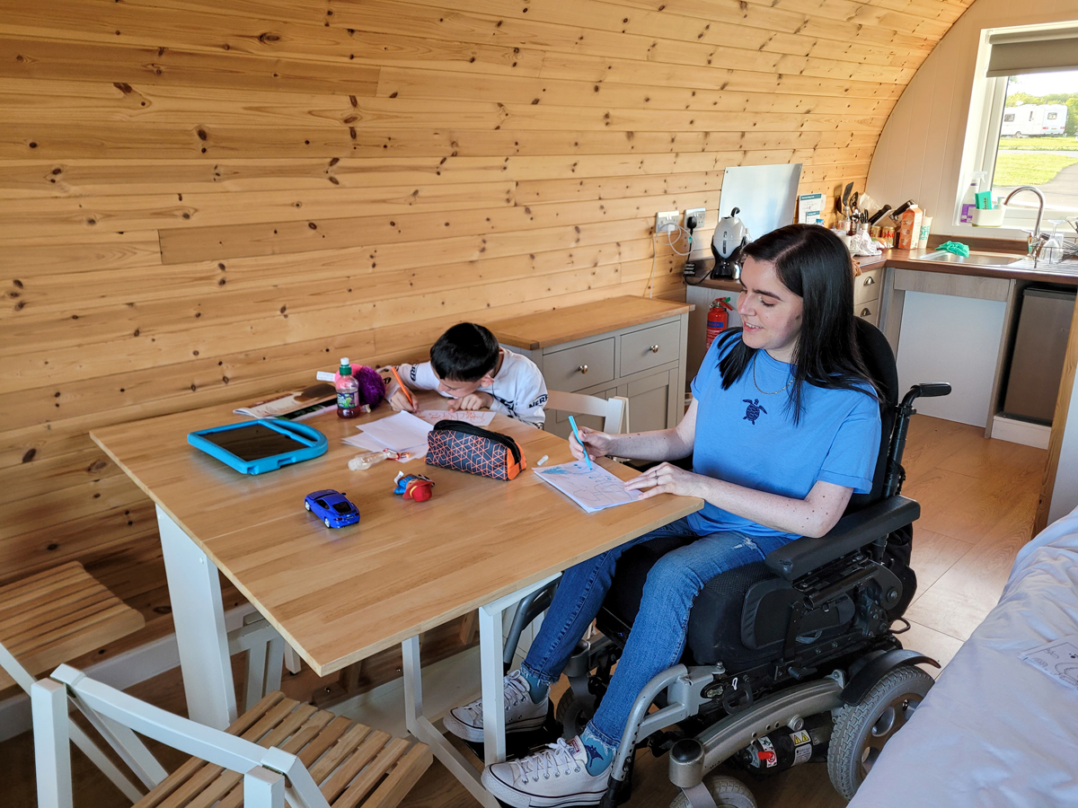 Emma and her nephew are sat at the dining table in the glamping pod. They are both drawing and colouring in.