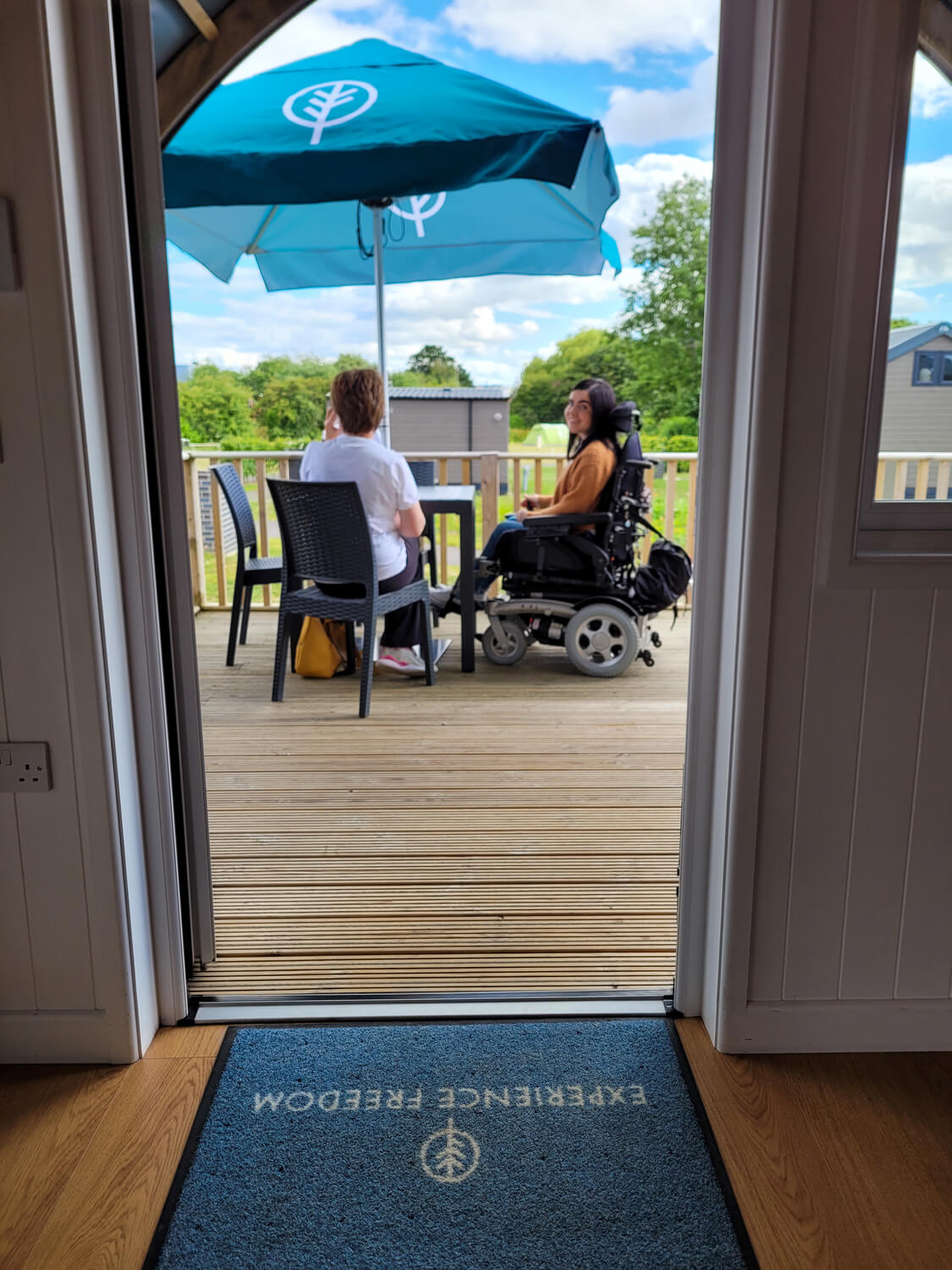 Emma sat at the outdoor dining table on the decking with her mum. A blue parasol is above them. Emma is looking over her shoulder and smiling at the camera.