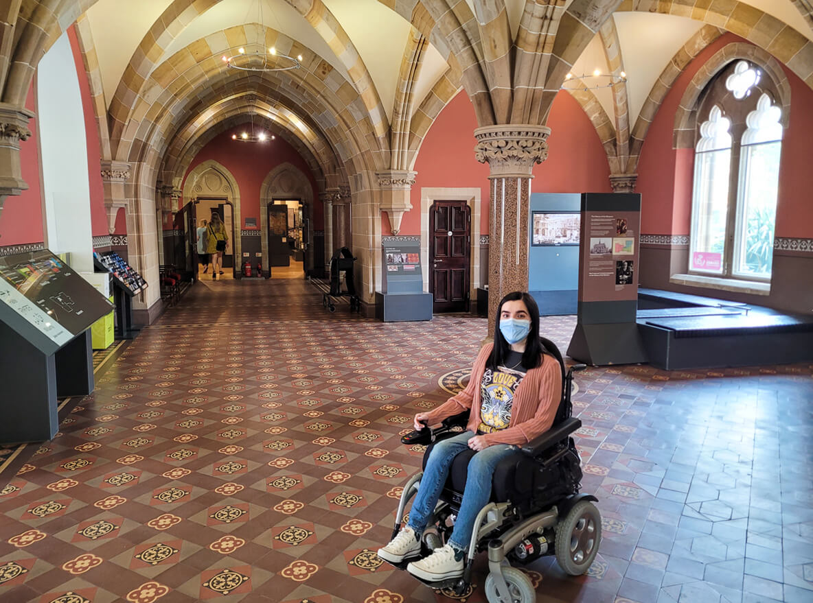 Emma sat in her wheelchair inside the McManus Museum in the reception area. The ceiling has gothic arches.