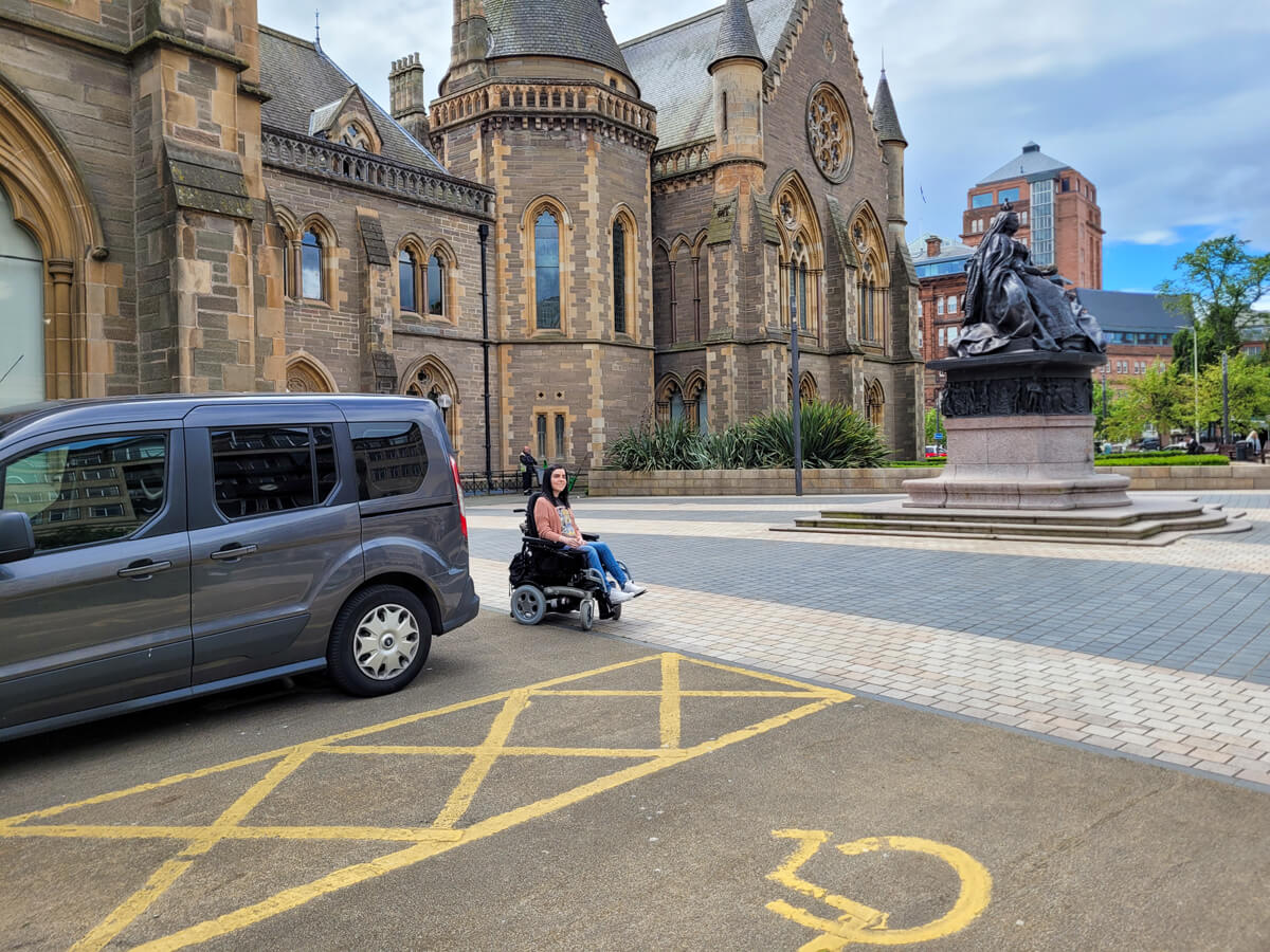Emma sat in her powered wheelchair at the rear of her wheelchair accessible vehicle (WAV). Her WAV is parked in a blue badge parking bay at the McManus Art Gallery & Museum Dundee.